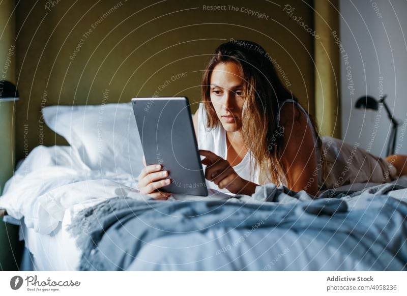Focused young woman browsing tablet while lying on bed in room using device home online social media surfing connection contemporary female modern comfort relax