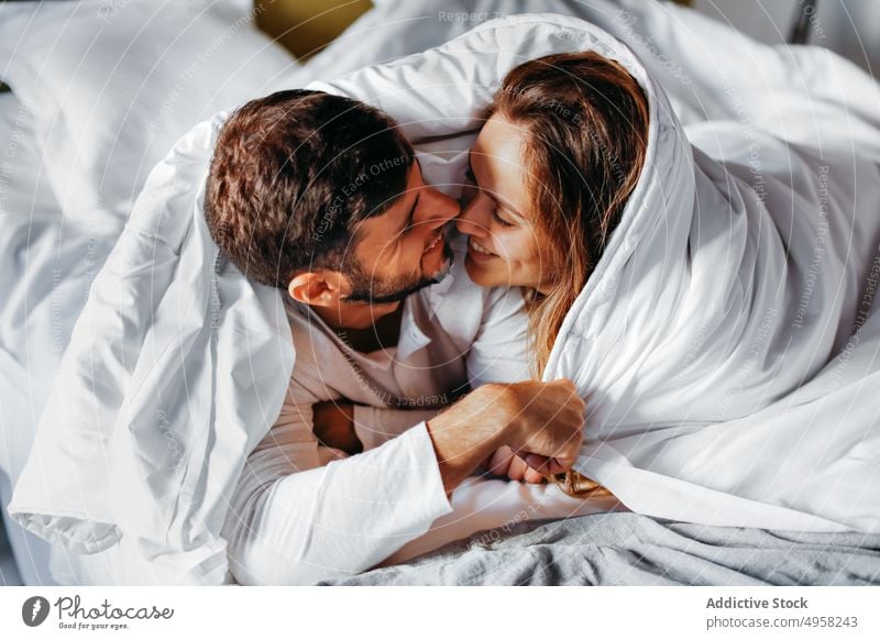 Cheerful young couple caressing under white blanket in bed embrace tender cuddle affection duvet together relationship happy love smile romantic bedroom wife