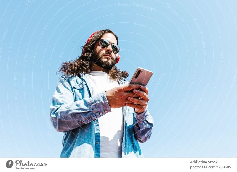 Ethnic man in sunglasses with smartphone in sunlight male style browsing individuality happy headphones lifestyle bearded using gadget device contemporary