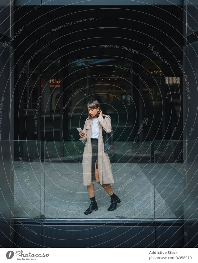 Woman with smartphone near modern building woman browsing online earphones tws earbuds urban street trendy outfit coat style fashion structure lifestyle