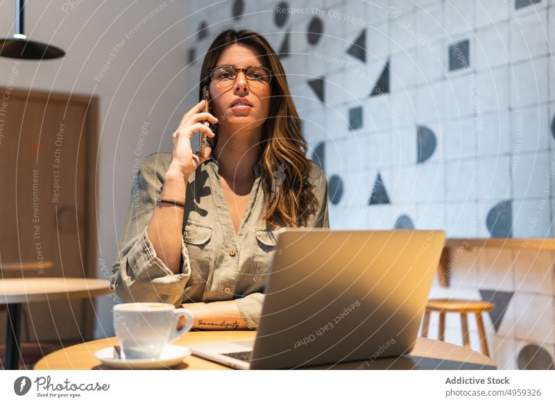 Woman speaking on smartphone against laptop in cafeteria woman talk hot drink busy work multitask table using gadget device cellphone beverage wall cup alone