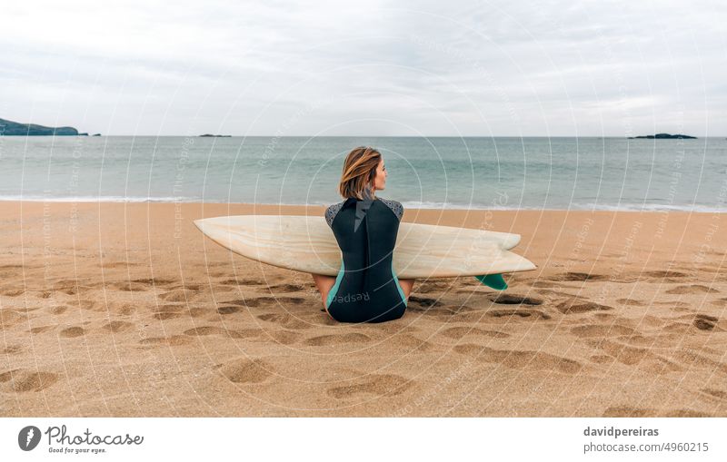 Surfer woman with wetsuit and surfboard sitting on the sand looking aside on the beach young surfer sea winter back people 20s outdoors twenties sport cold wave