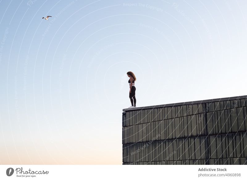 Anonymous sportswoman resting on roof under light sky sportswear freedom admire vitality energy sunset stand enjoy building wall contemplate sundown