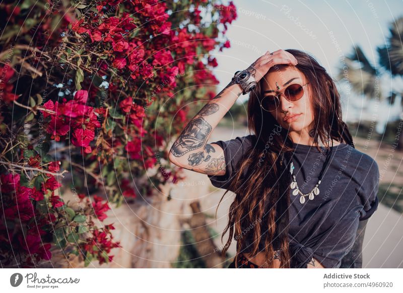 Woman in sunglasses against bush with colorful flowers in park woman cool tattoo rebel allure piercing bindi appearance brunette eye gaze trendy tradition