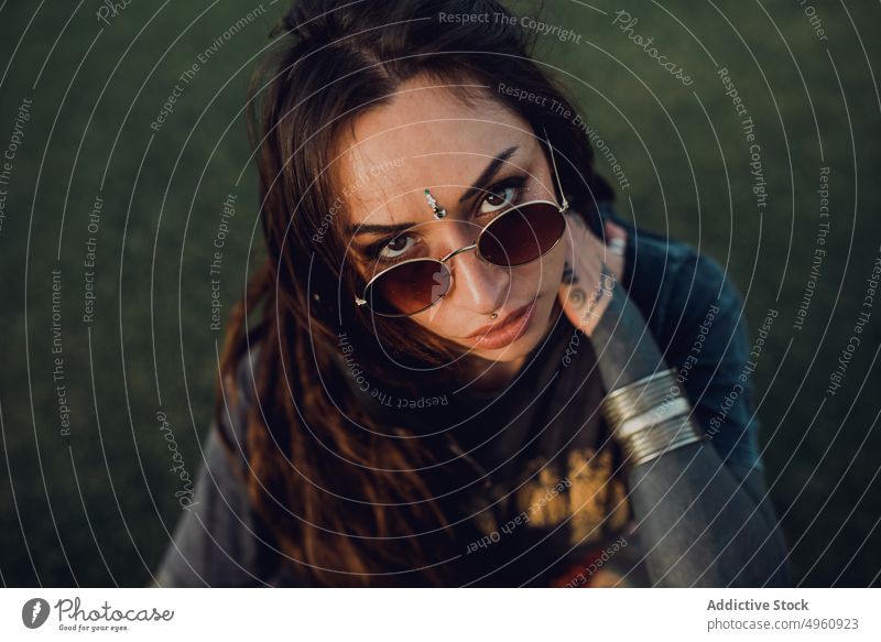 Calm funky millennial woman on lawn in park tattoo allure appearance brunette jewelry sunglasses cool piercing calm accessory provocative grass fashion female