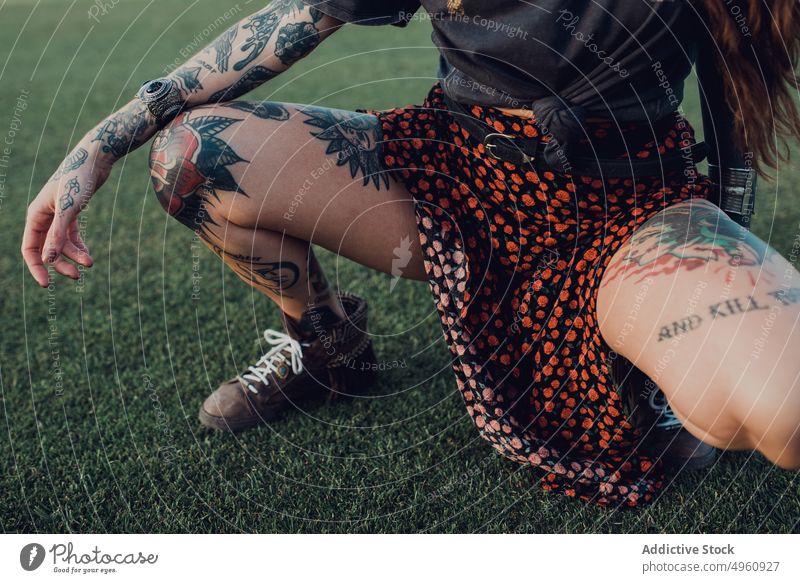 Stylish millennial tattooed woman on lawn brunette leg cool rebel trendy provocative squatted fashion female style gorgeous individuality sneakers classy vogue
