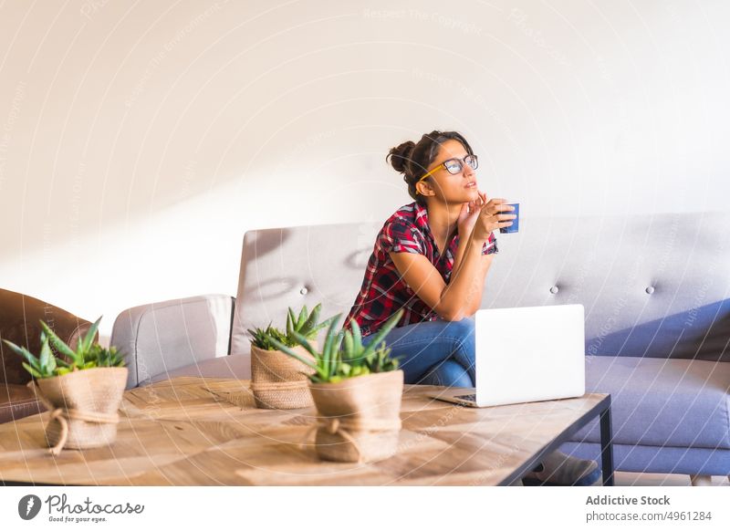 Thoughtful woman on sofa against assorted cacti in hotel pensive touch chin plant tropical exotic table thoughtful checkered ornament denim wear sit eyewear bun