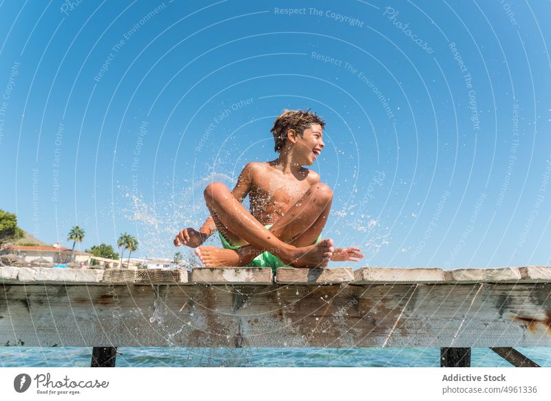 Cheerful teenager splashing water on pier boy summer cheerful sea vacation having fun holiday smile sunny weekend sit wooden quay wet hair delight content