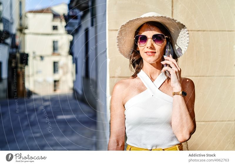 Positive woman talking on smartphone in city conversation phone call street summer hat speak mobile gadget sunglasses female happy device enjoy content cheerful