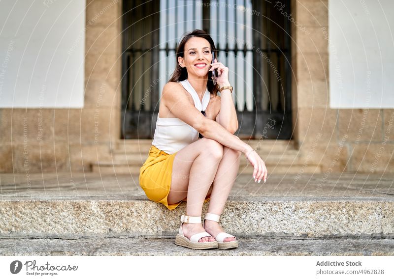 Happy young lady having phone call sitting on step on street woman talk smartphone smile conversation happy communicate optimist mobile female dark hair style