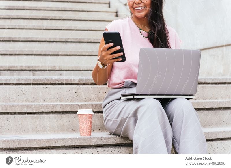 Crop ethnic woman sitting on stairs while using laptop and smartphone on street freelance smile remote takeaway coffee work online connection female young