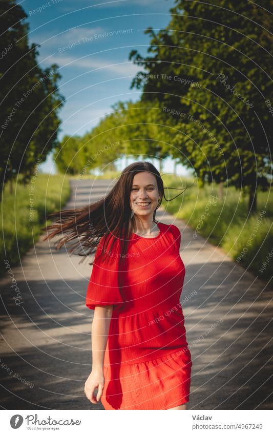 Beautiful young brunette in a red summer dress with flowing brown hair walks along a bicycle path and in an avenue of trees on a summer day. Candid portrait of a student