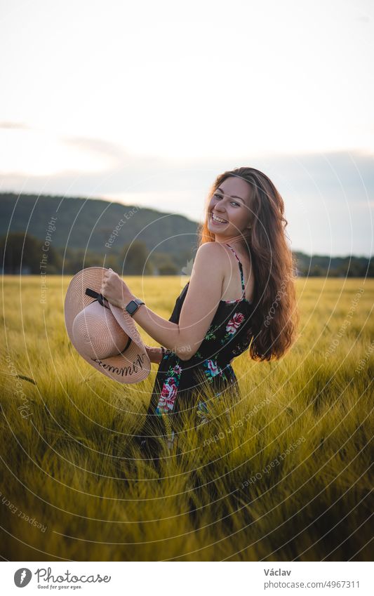 Wonderful natural smile of a full grown lady in a flowered dress with brown hair in the middle of a meadow wearing a beautiful wicker hat. Candid portrait of young model at sunset