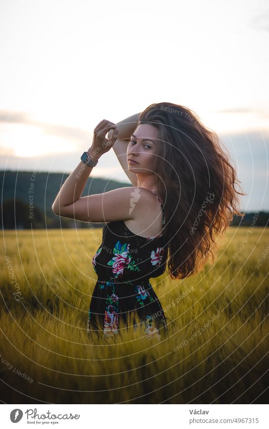 Beautiful realistic smile of brunette in black dress standing in the middle of a field during sunset. Candid portrait of inner emotions. Ruffle your hair.