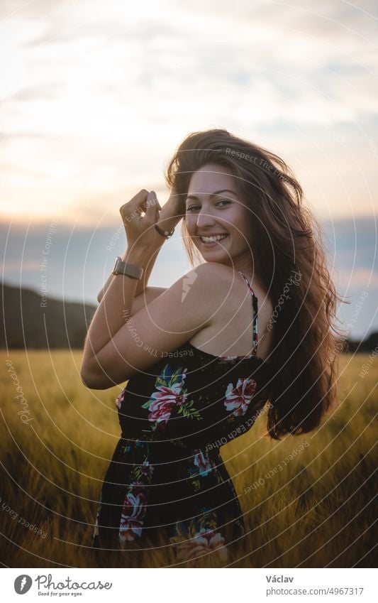Beautiful realistic smile of brunette in black dress standing in the middle of a field during sunset. Candid portrait of inner emotions. Ruffle your hair summer