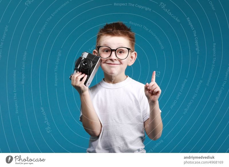 Little boy taking a picture using a retro camera and with finger pointed up. Child boy with vintage photo camera isolated on blue background. Old technology concept. Child learning photography