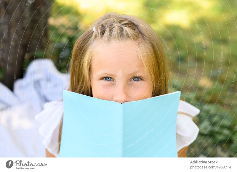 Girl with book in park girl cover face summer weekend story daytime free time blond gray eyes season childhood nature kid literature read notebook notepad