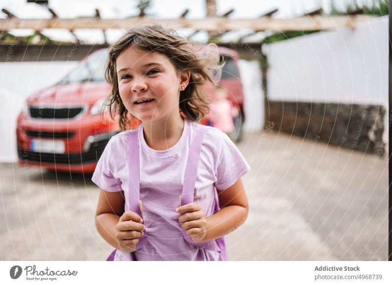 Girl with backpack near car in yard girl smile weekend summer travel ready happy child backyard casual daytime vehicle glad carefree cheerful positive optimist