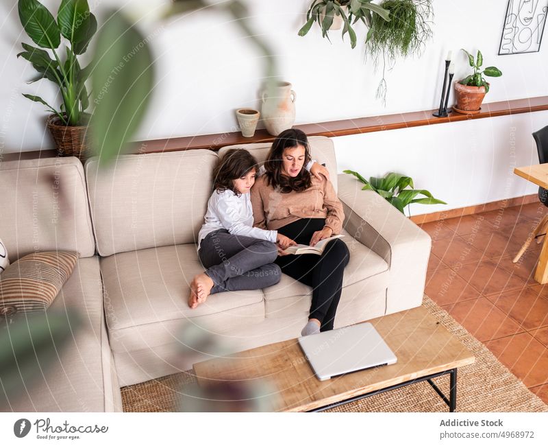 Woman reading book with daughter woman girl sofa mother home living room child together female couch plant hobby literature motherhood apartment kid parent