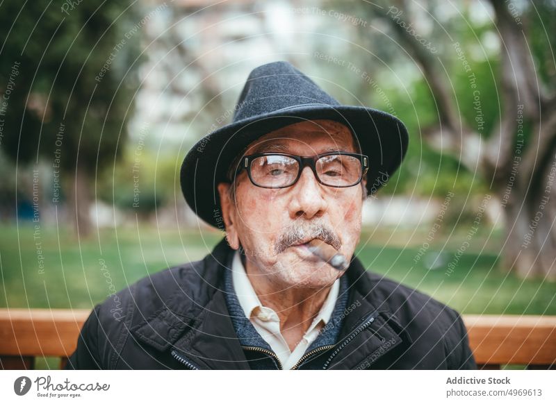 Elderly man smoking cigar on street senior elderly aged smoke park habit nicotine bench hat casual lifestyle city mustache pensive headwear headdress outfit