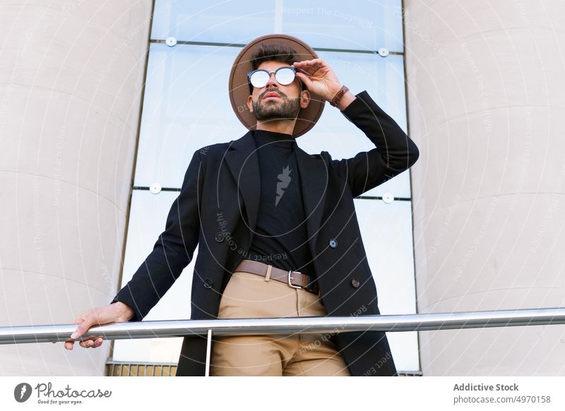 Masculine man in trendy sunglasses behind handrail in town fashion vogue style individuality creative design contemporary masculine hat jacket macho stylish