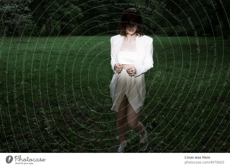 Dark woods and a green meadow. A fashion model is running into the spotlight. Dressed in a white avant-garde outfit she is looking splendidly. A moody atmosphere in this action shot.