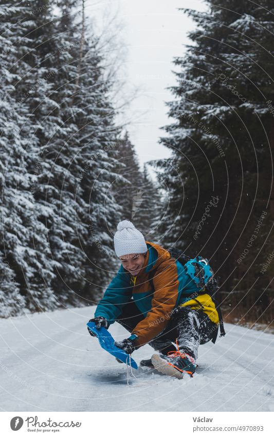 Candid portrait of a young man wearing a colorful winter hiking jacket, warm-ups and a white cap during the winter season in a snowy area. Riding a plastic snowboard