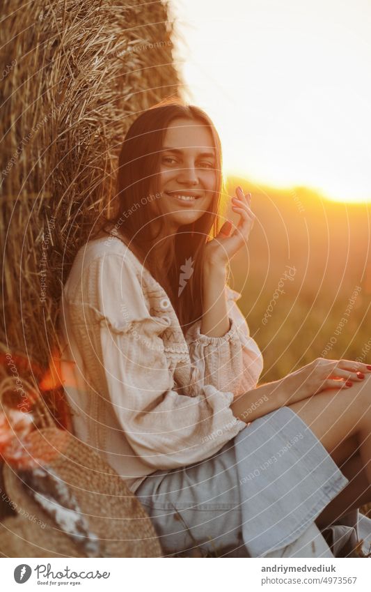 young woman in the beautiful light of the summer sunset in a field is sitting near the straw bales. beautiful romantic girl with long hair outdoors in field
