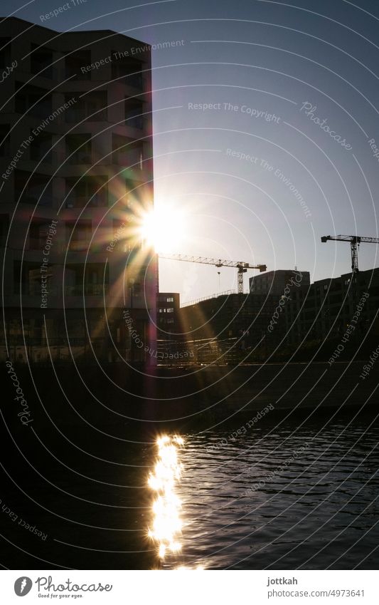 Sun rays behind a modern multi-storey house, construction cranes next to it, water in the foreground Berlin Construction site Building Architecture