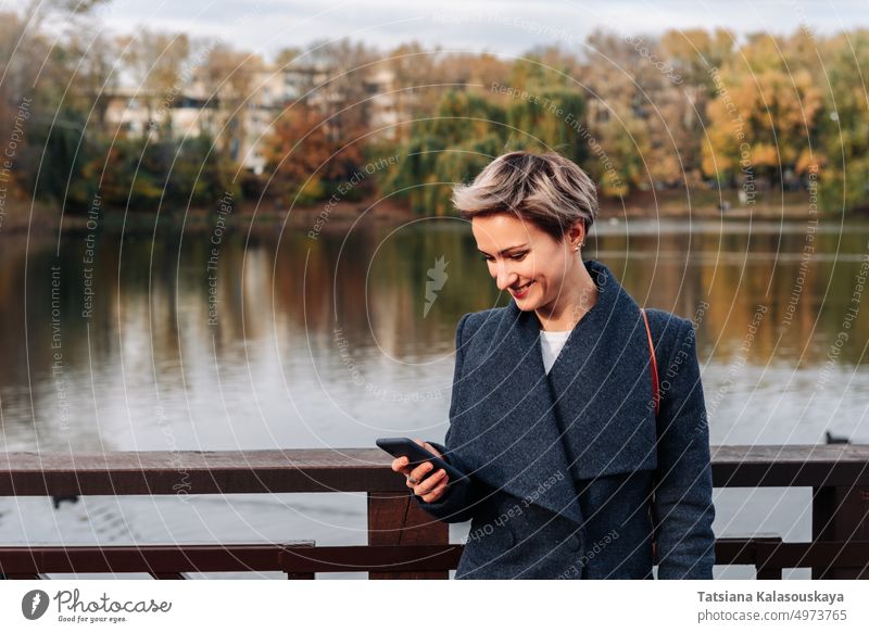 A woman in a city park by the river uses a smartphone female adult short hair Blond Attractive cell cell phone happiness happy joy smile Lifestyles real people