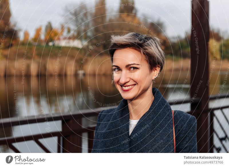 A woman smiles broadly near the river in autumn fall short-haired Lifestyle female happiness adult happy joy 30-35 years cheer cheerful city park laughter