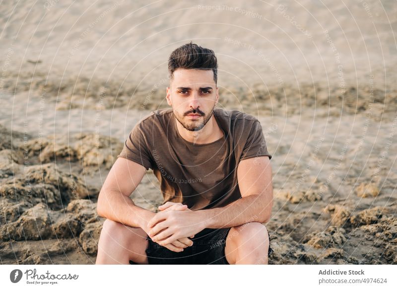 Handsome young Hispanic man sitting on sandy beach clasping hands thoughtful contemplative brunet resting folding relaxing ethnic hispanic waiting thinking