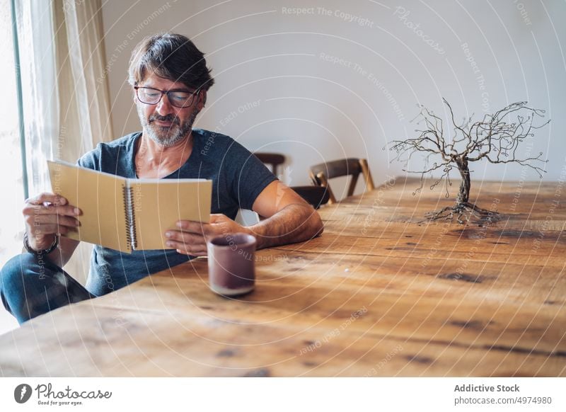 Focused aged man reading notebook at home focused calm content concentration sit table wooden cup checking serious senior beard glasses retired relax coffee tea