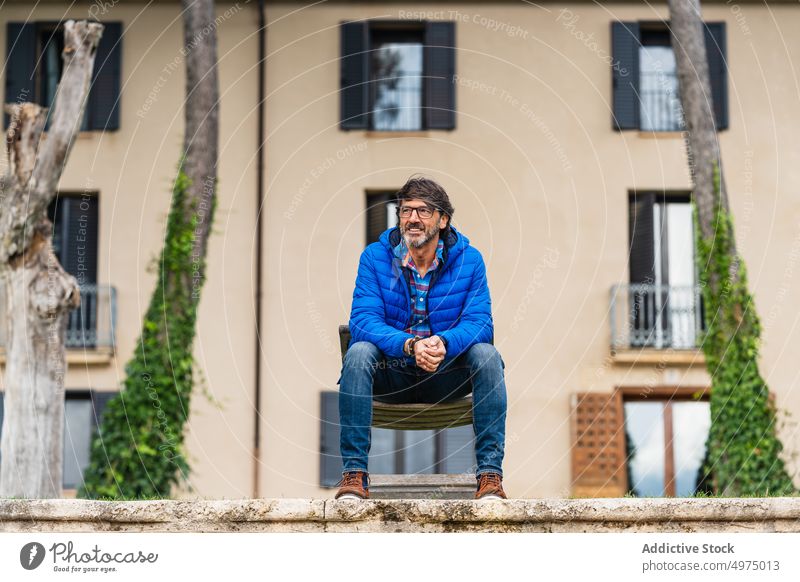 Middle-aged man sitting on a wooden chair while pensively leaning portrait blue wearing jacket one bearded stone column green sky day outdoor park winter