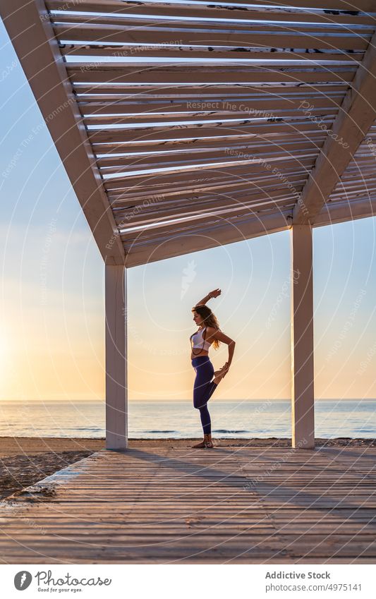 Woman doing yoga in standing split pose at seaside woman balance flexible sportswear seashore sunrise practice healthy harmony wellness slim stretch posture