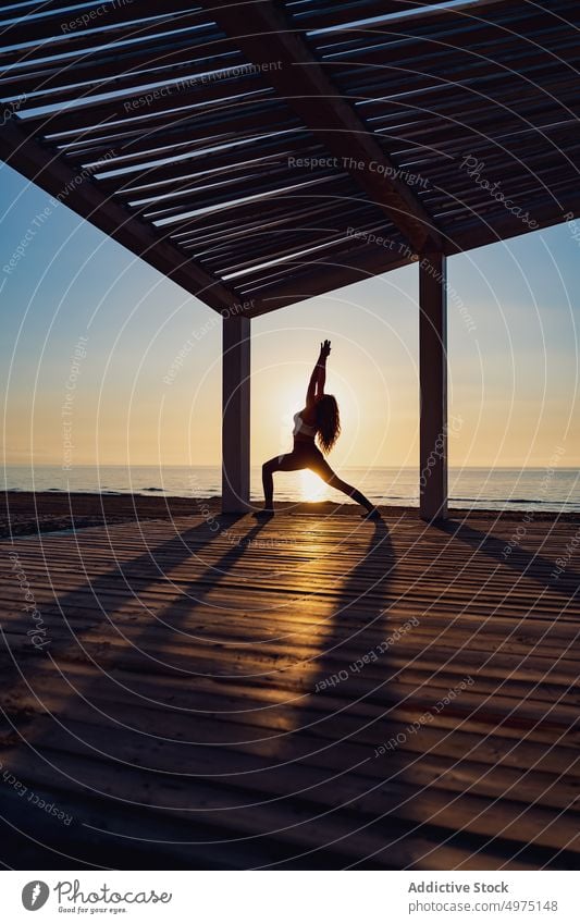 Tranquil woman doing yoga in Warrior pose during sunrise warrior seaside practice tranquil silhouette dawn wooden terrace healthy harmony relax asana