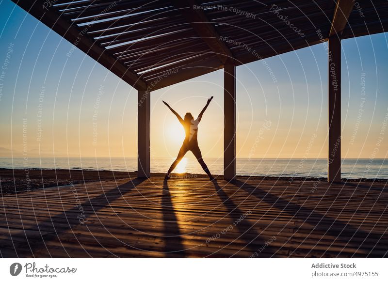 Tranquil woman doing yoga with arms and legs open during sunrise pose seaside practice tranquil silhouette dawn female wooden terrace healthy harmony warrior