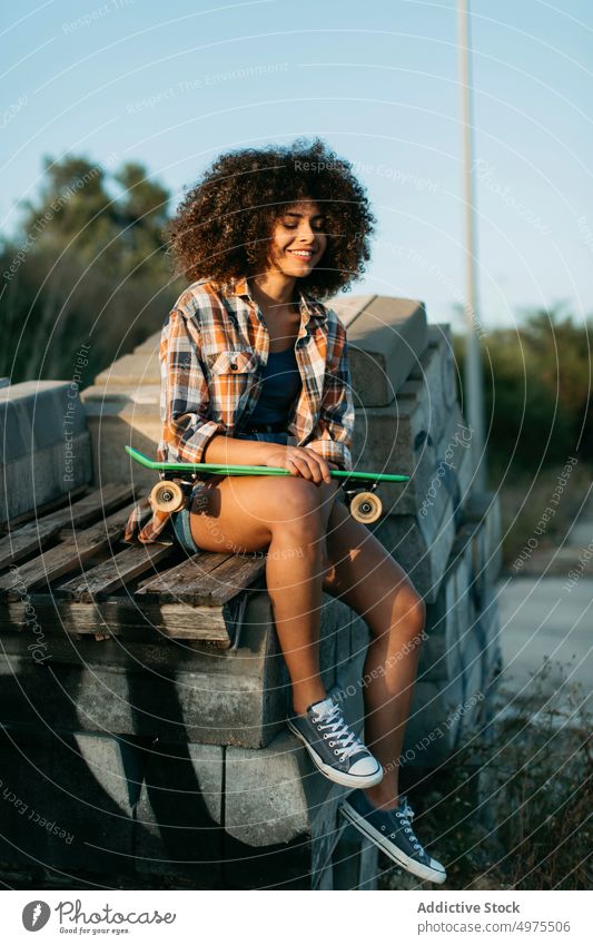 African American woman with penny board sitting on stones on the street summer skater cool millennial afro trendy tranquil hipster sunny female ethnic