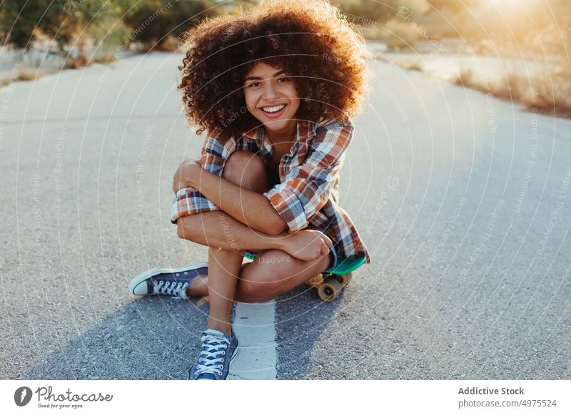 Delight African American woman sitting on penny board on the asphalt road summer skater street cool millennial afro trendy tranquil hipster sunny female ethnic