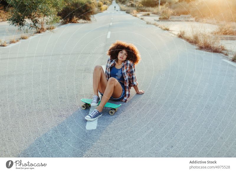 African American woman sitting on penny board on the asphalt road summer skater street cool millennial afro trendy tranquil hipster sunny female ethnic