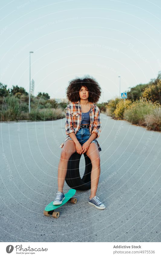 Delight African American woman sitting on tire with penny board on the asphalt road summer skater street cool millennial afro trendy tranquil hipster sunny