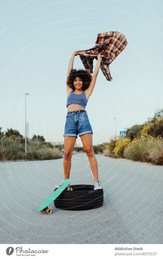 Delight African American woman sitting on tire with penny board on the asphalt road summer skater street cool millennial afro trendy tranquil hipster sunny