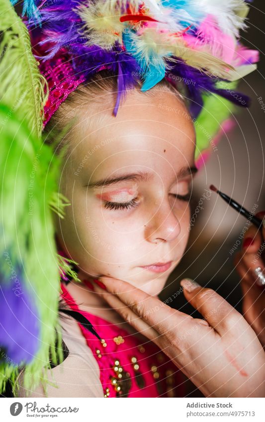 Crop mother applying festival makeup on daughter face carnival prepare airliner home colorful feather headgear girl woman kid child fashion event glamour
