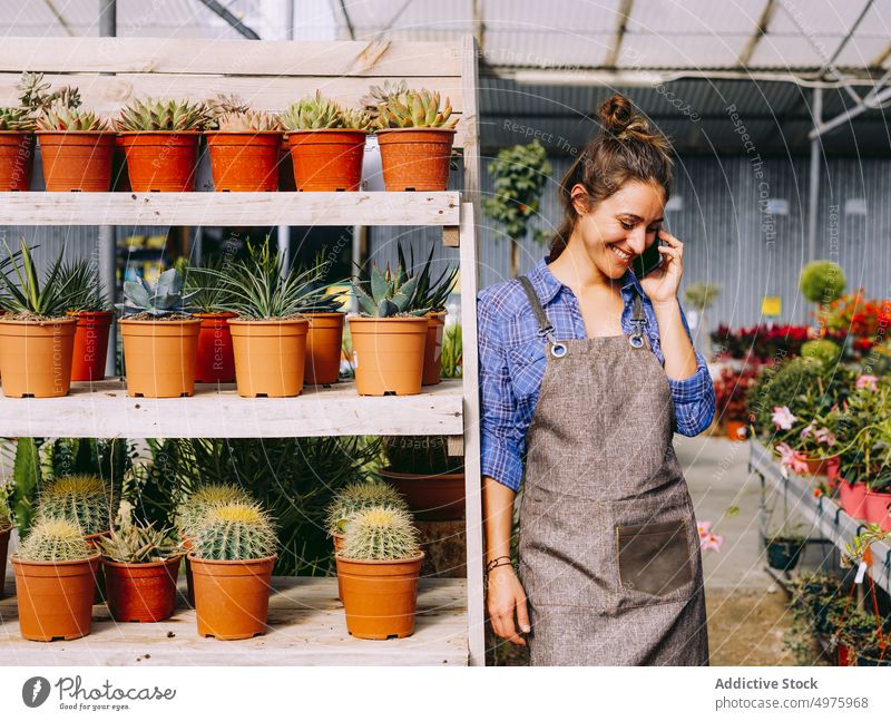 Cheerful florist speaking on smartphone in greenhouse gardener talk woman smile lean shelf work organic botany female small business plant professional