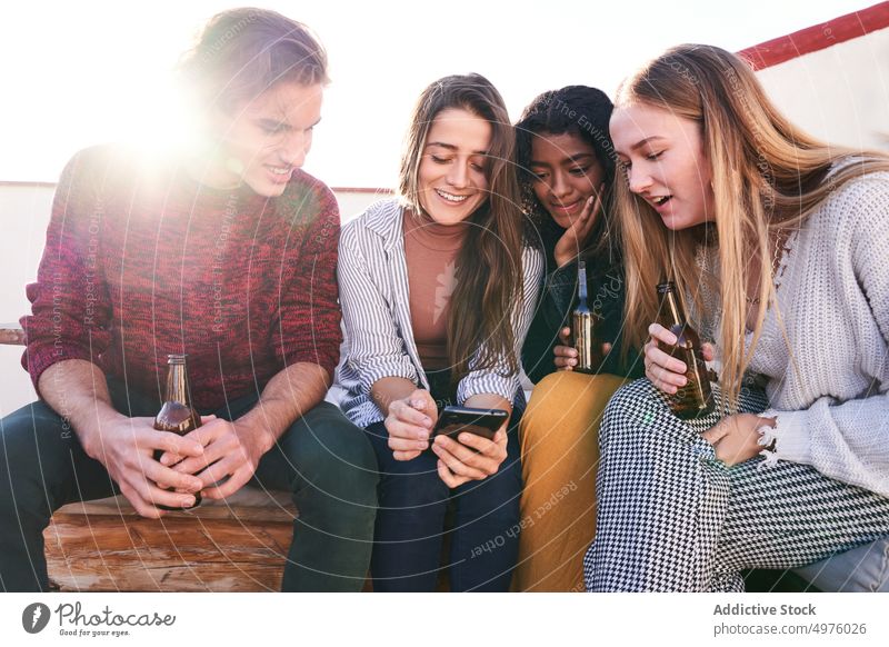 Cheerful friends with beer using smartphone on sofa on terrace content party cheerful booze together device gadget watch happy friendship share gather surfing