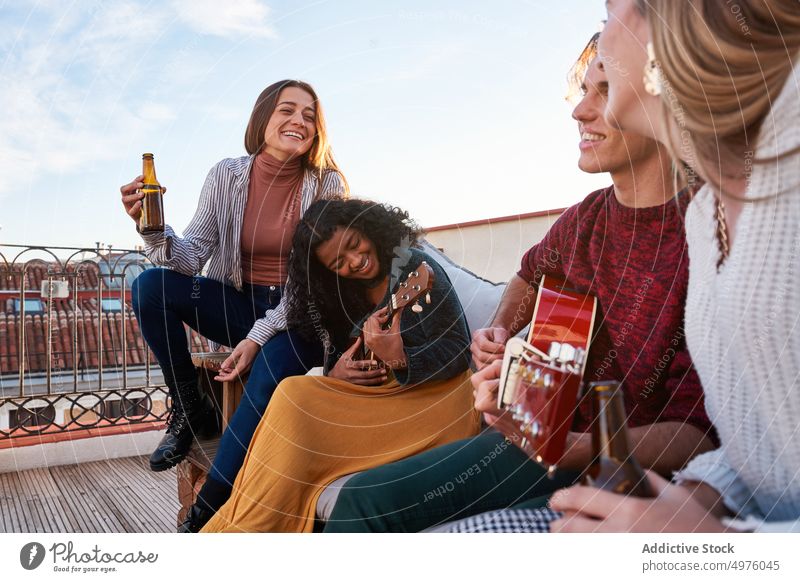 Joyful diverse friends playing guitar and ukulele on rooftop terrace content melody music sofa instrument cheerful together guitarist sound friendship gather