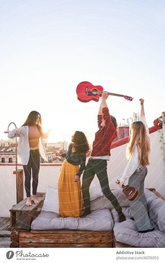 Delighted multiethnic friends playing guitar and jumping on terrace sofa rooftop excited music instrument having fun party merry together group cheerful