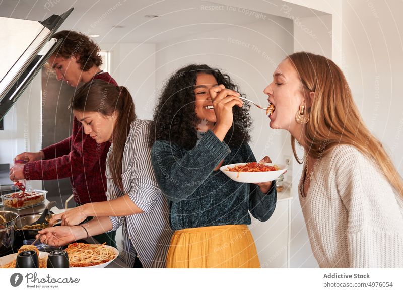 Cheerful ethnic woman spoon feeding friend with delicious pasta cheerful spaghetti happy care food kitchen appetite eat relationship hungry together meal dinner