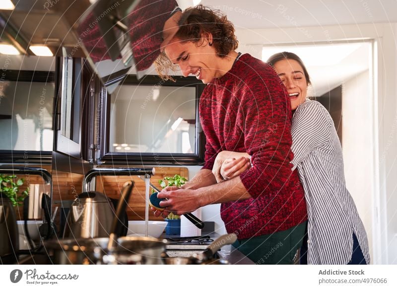 Joyful woman hugging boyfriend while cooking together in kitchen couple delight wash sink toothy smile embrace domestic relationship happy lifestyle chore