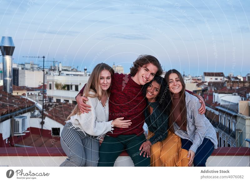 Smiling multiracial friends hugging and sitting on rooftop in twilight toothy smile cheerful together embrace carefree gather young terrace veranda town city